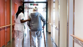 Photo d'un couloir d'hôpital. Une infirmière soutien une personne en train de marcher avec un déambulateur. Les deux personnes sont vues de dos. 