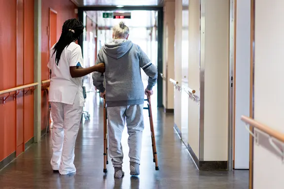Photo d'un couloir d'hôpital. Une infirmière soutien une personne en train de marcher avec un déambulateur. Les deux personnes sont vues de dos.