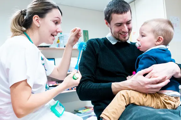Un papa enlace tendrement son fils tandis que le médecin essaye de détourner l'attention de l'enfant en jouant avec un palmier en plastique.